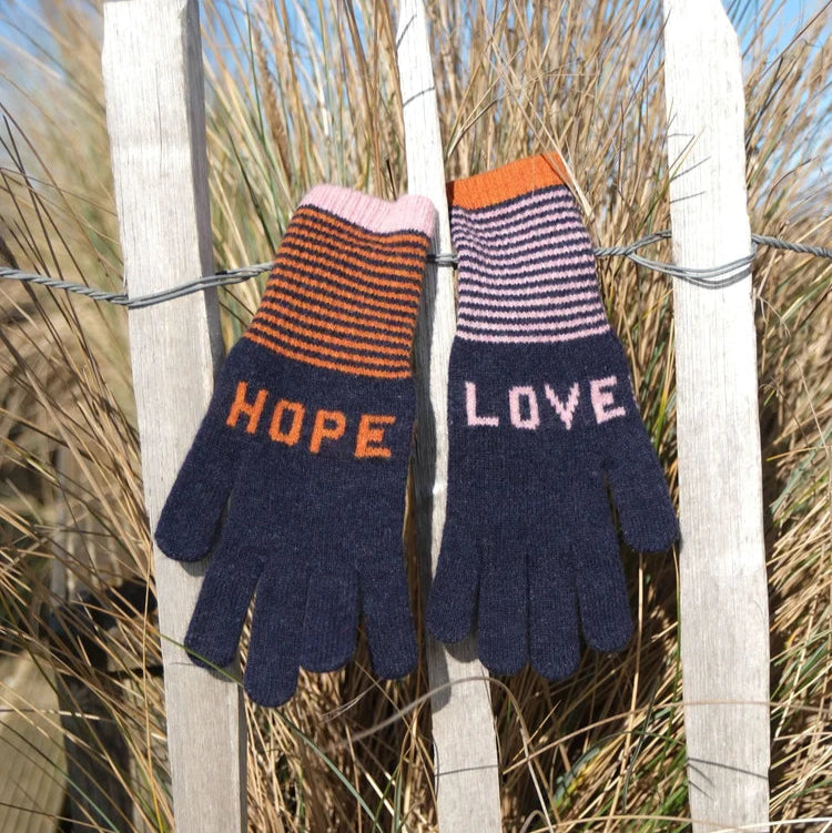 Pair of knitted gloves with 'HOPE' and 'LOVE' text on a wooden fence with grass in the background