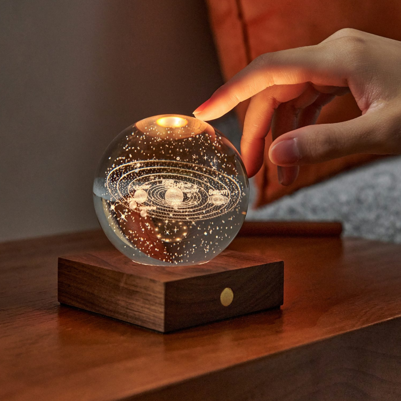 A hand touching a solar system motif amber crystal light on a walnut wood base, creating a warm glow.