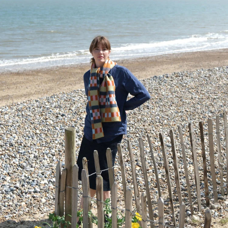 Person standing on a pebble beach with ocean view, wearing a colourful scarf and blue sweater.