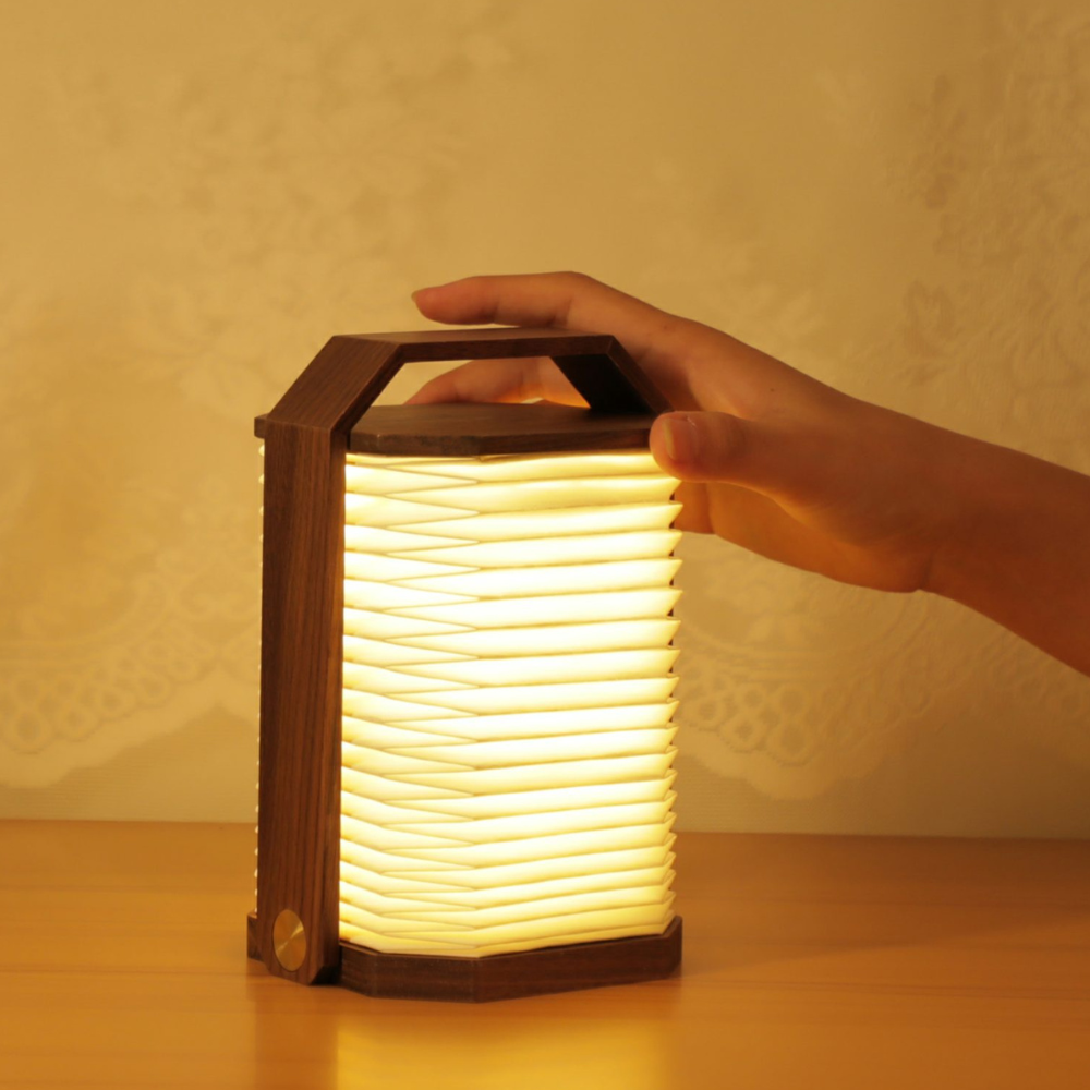 An illuminated origami walnut lamp with a hand holding it, displaying the glowing LED light through the folded paper.