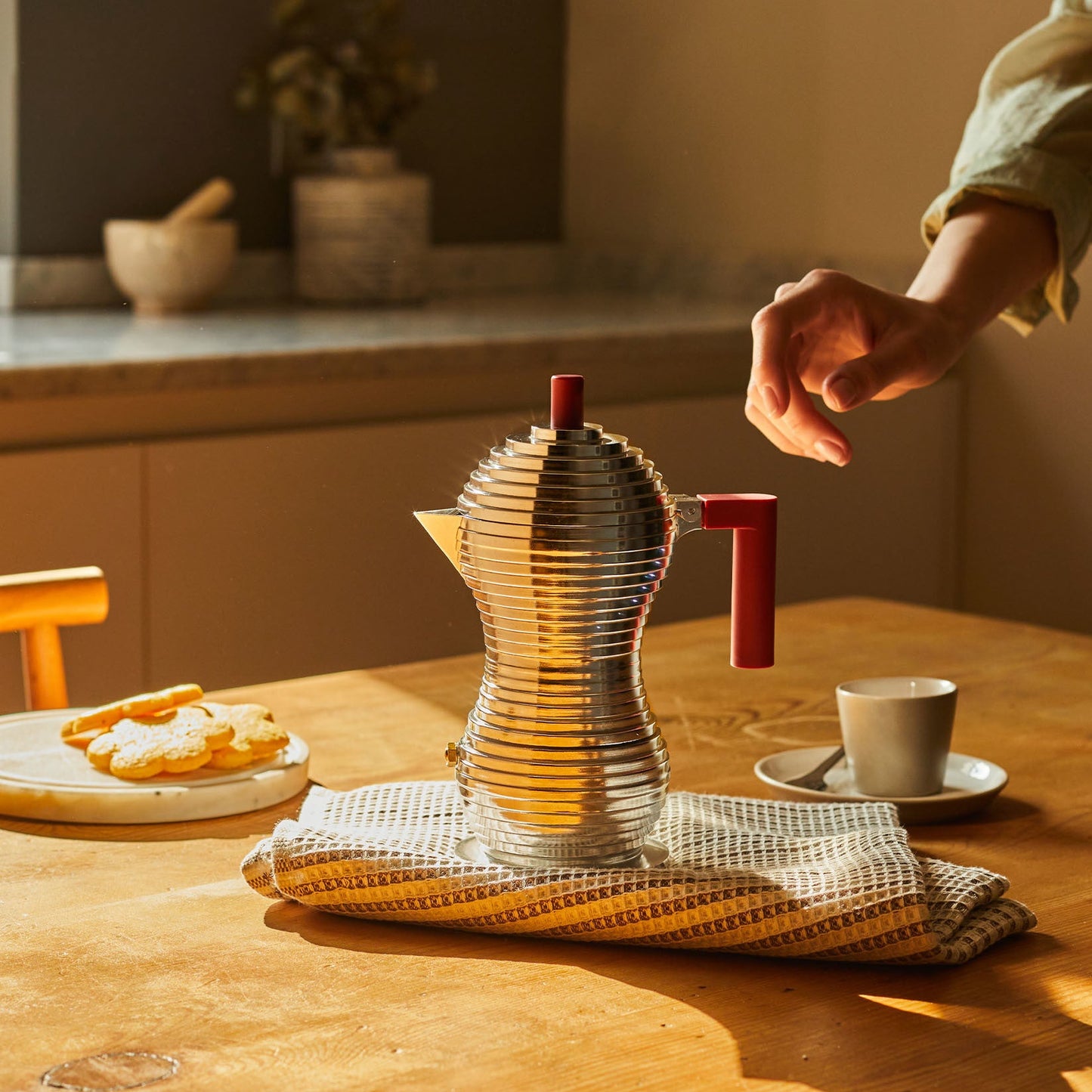 Person interacting with a unique teapot on a wooden table with a warm, homey atmosphere.