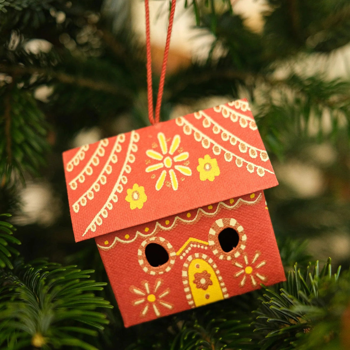 Decorative paper house ornament with yellow details hanging on a Christmas tree.