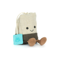 A beige and grey plush teabag, with a blue label, a smiling face, and two legs.