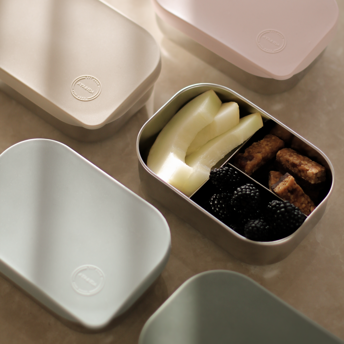 Four minimalist rectangular lunch boxes in soft pastel colours are arranged on a warm beige surface. One box is open and contains sliced white fruit and brown snack pieces, while the others are closed with their lids resting beside them.