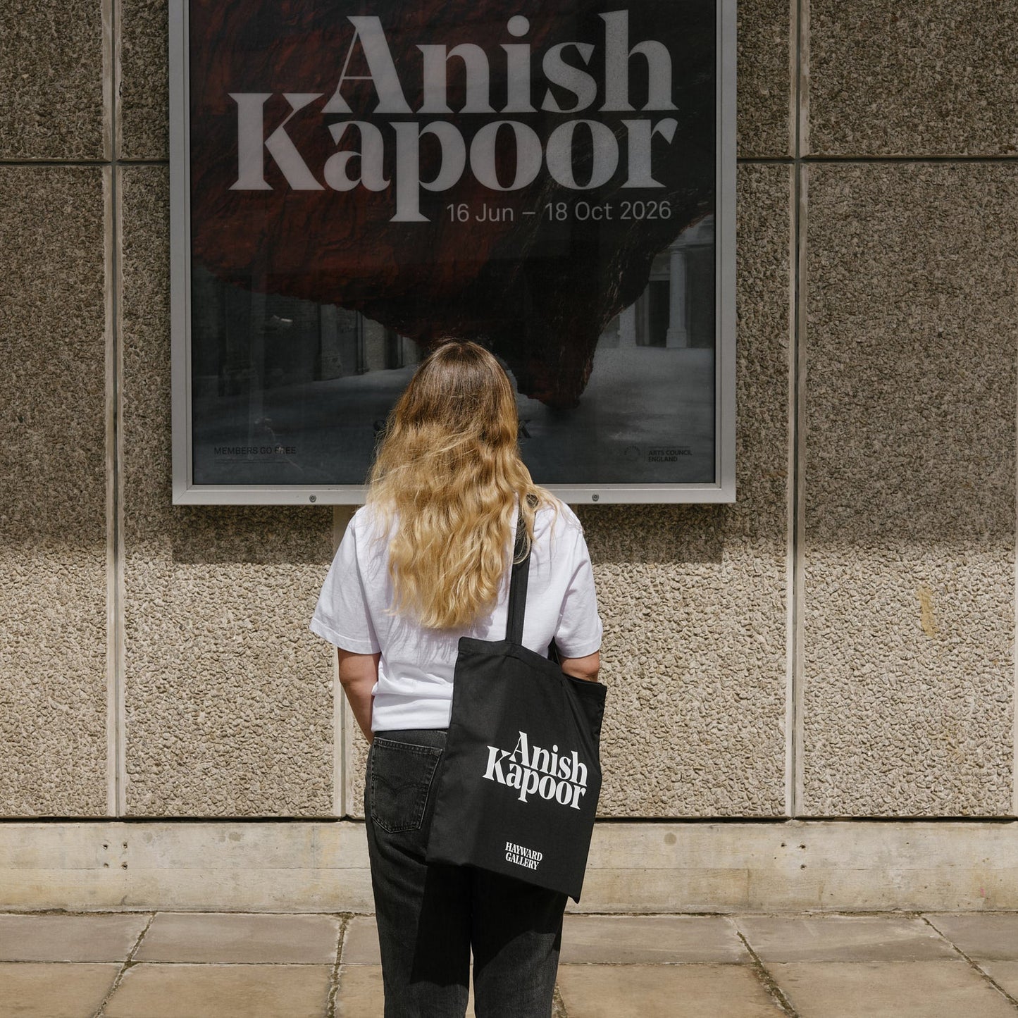 Person with a 'Anish Kapoor' tote bag looking at an art exhibition poster on a wall.