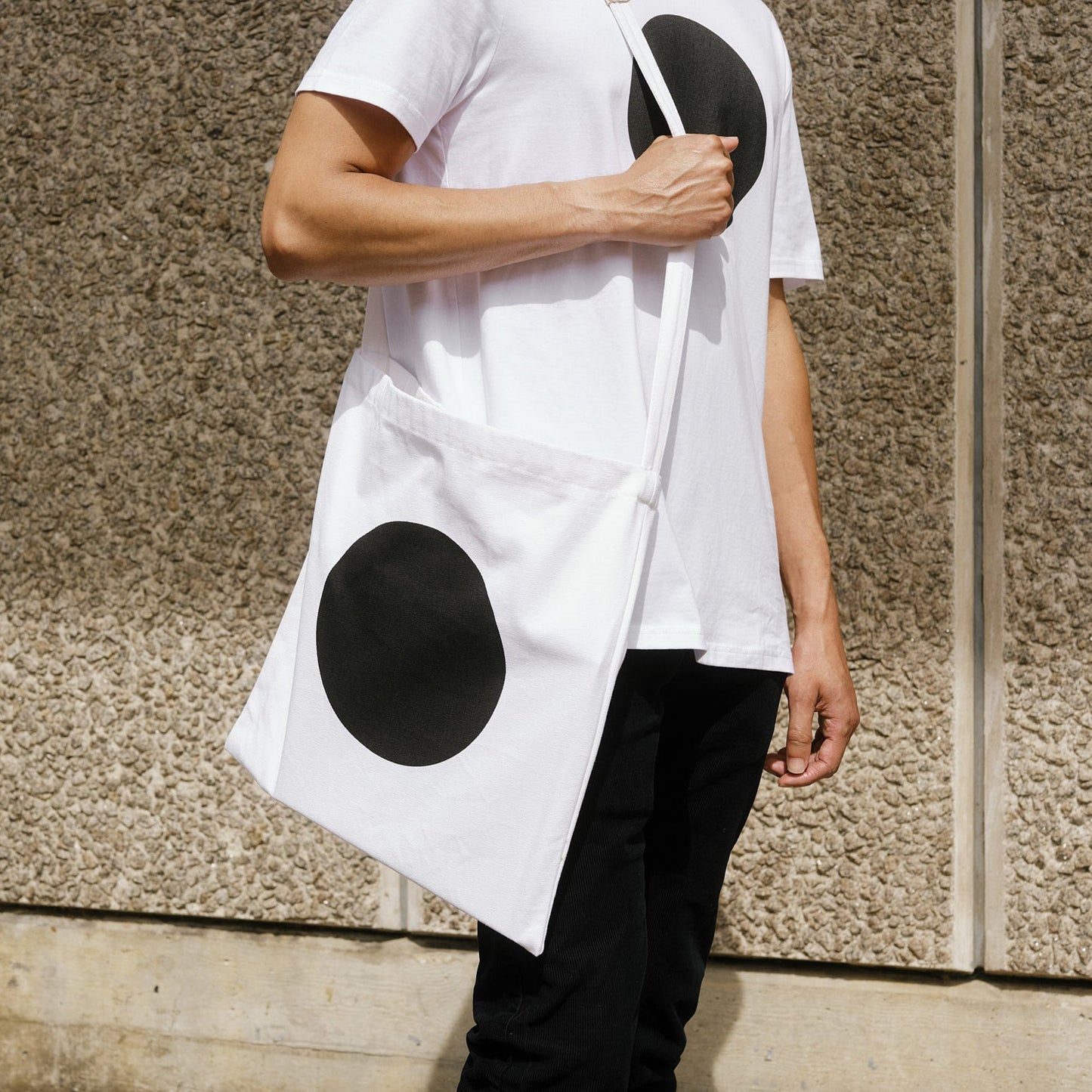 Person wearing a white shirt and tote bag with black circular patterns against a stone wall.