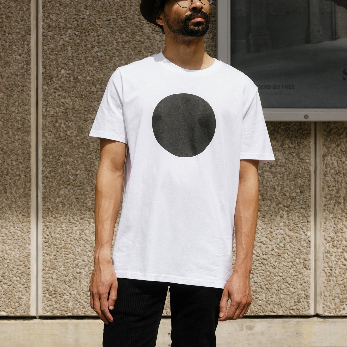 Man wearing a white t-shirt with a black circle design, standing against a textured wall.