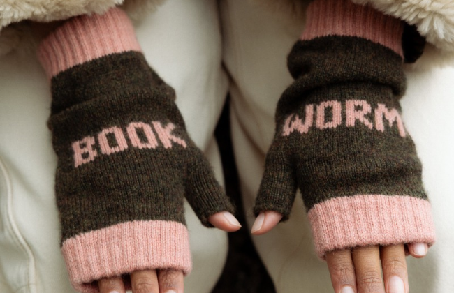 A close-up of a person wearing dark brown knit fingerless gloves with pink ribbed cuffs and knitted lettering that reads 'BOOK' on one glove and 'WORM' on the other.