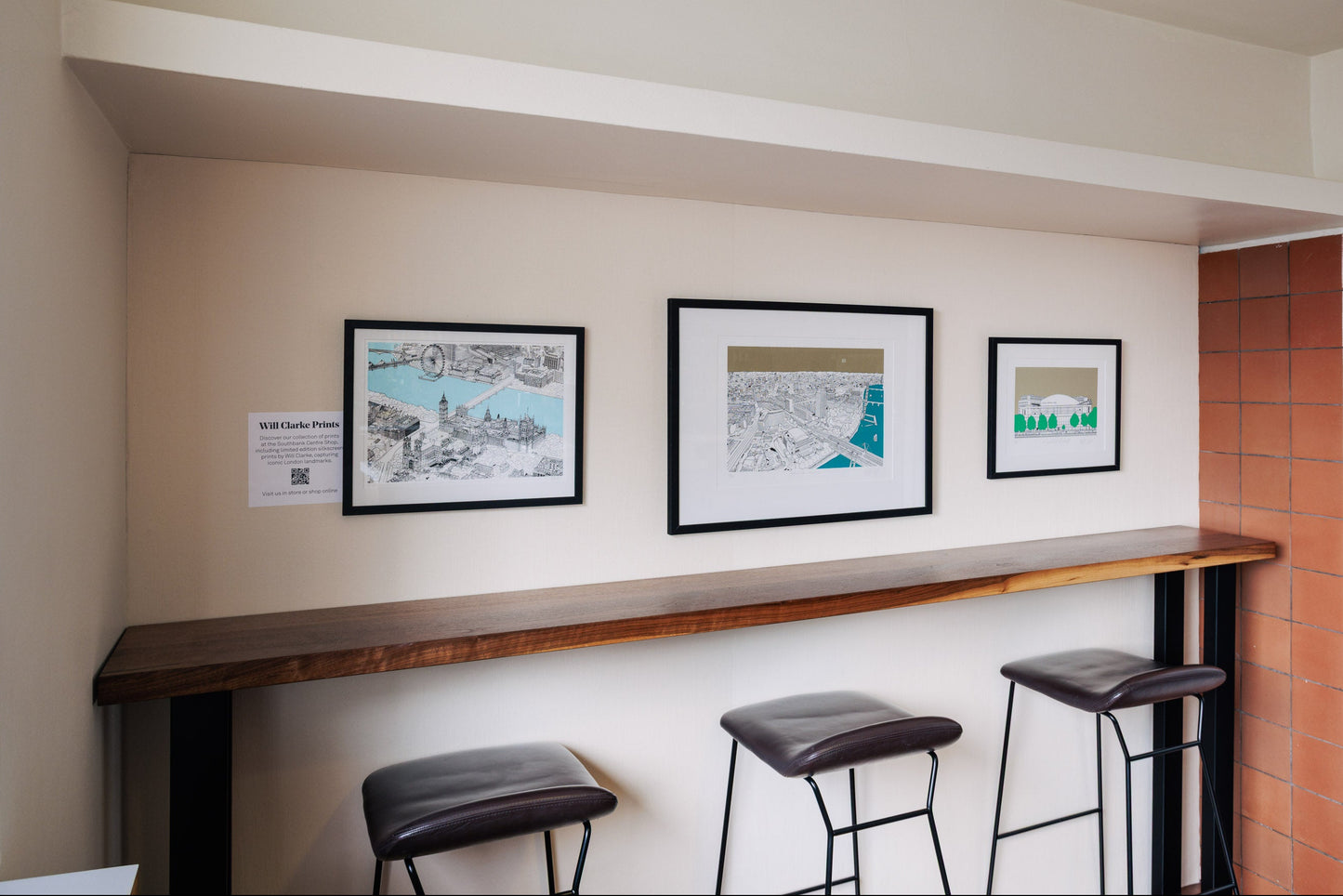 Three framed London scene artworks on a wall above bar stools in a room with tiled walls.