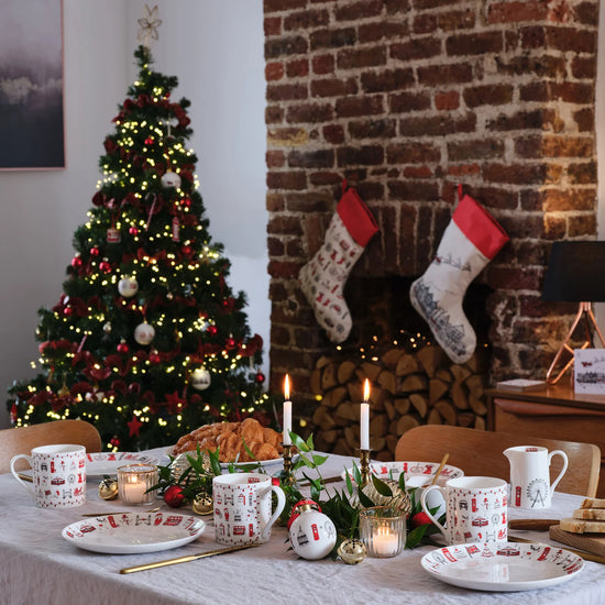 Table top decorated with London themed tableware including mugs, plates and baubles. A Christmas tree and a fireplace with two stockings hanging in the background.