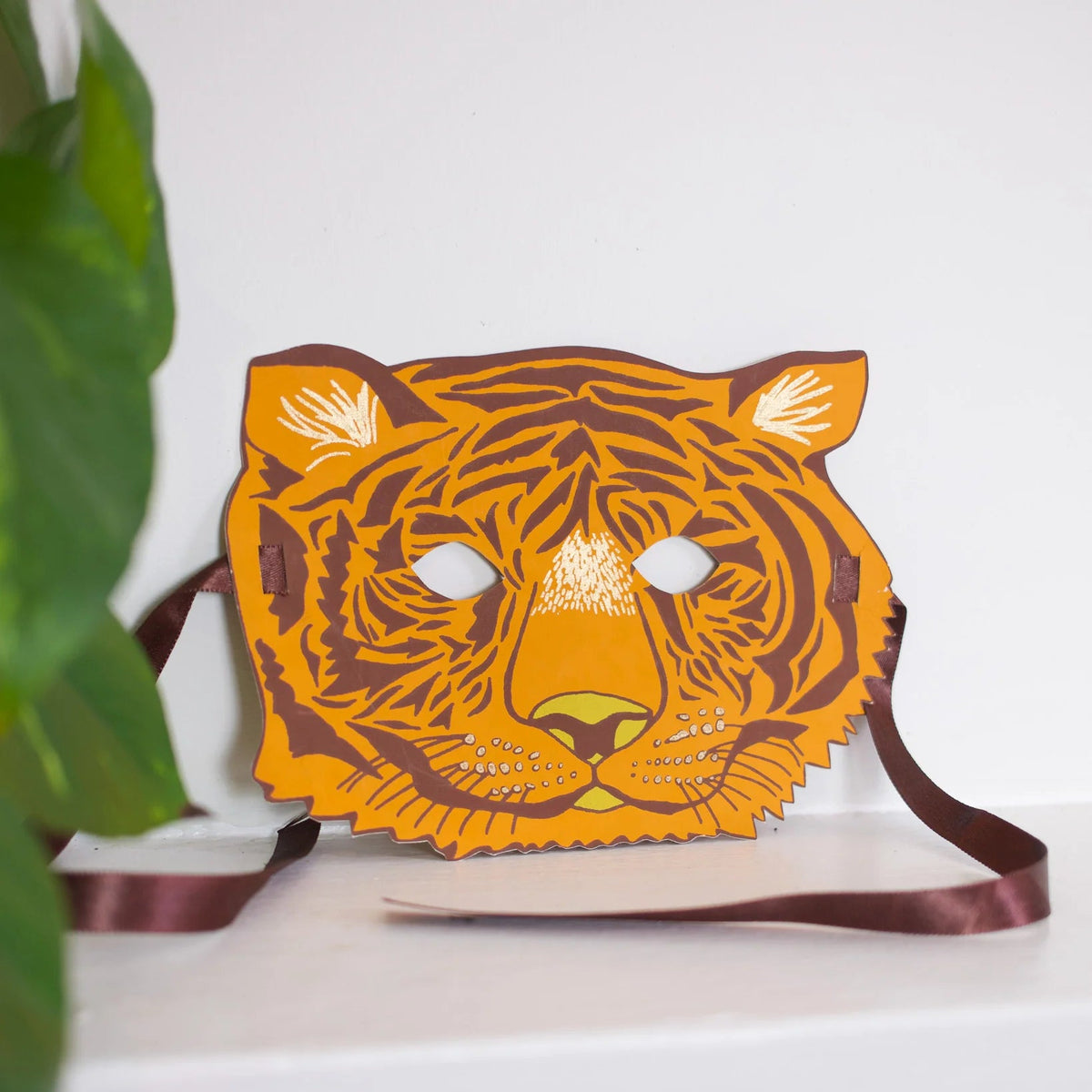A tiger head mask with a brown ribbon strap, displayed on a white shelf next to a green plant leaf against a white wall.