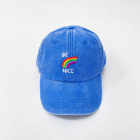 A vintage blue denim cap with 'BE NICE' rainbow artwork by David Shrigley embroidery on the front.