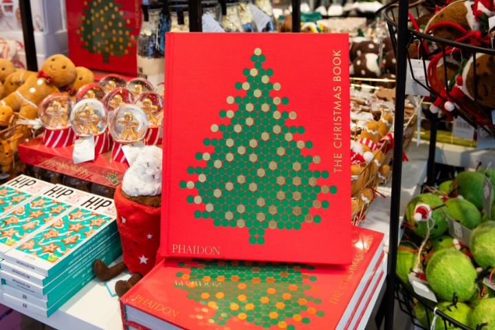 The Christmas Book displayed on a stack of books, on a table with other Christmas gifting products including a gingerbread snow globe and gingerbread decorations.