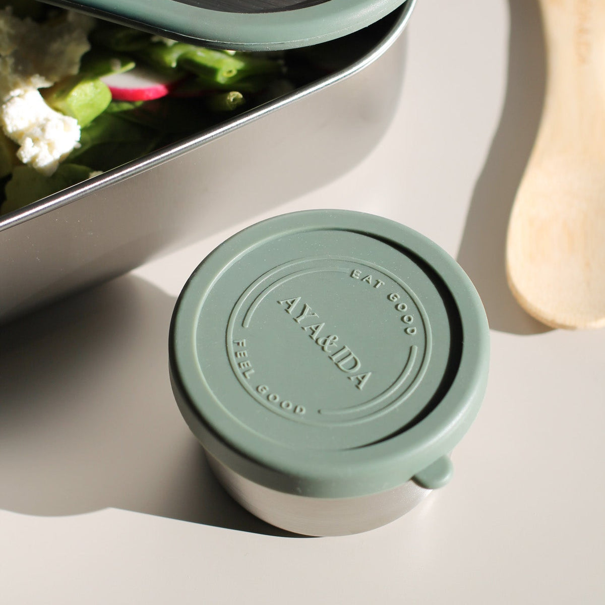 A close-up of a small round reusable container with a muted sage green lid and a white base, shown beside a metal lunch box with food and a wooden utensil on a light surface.