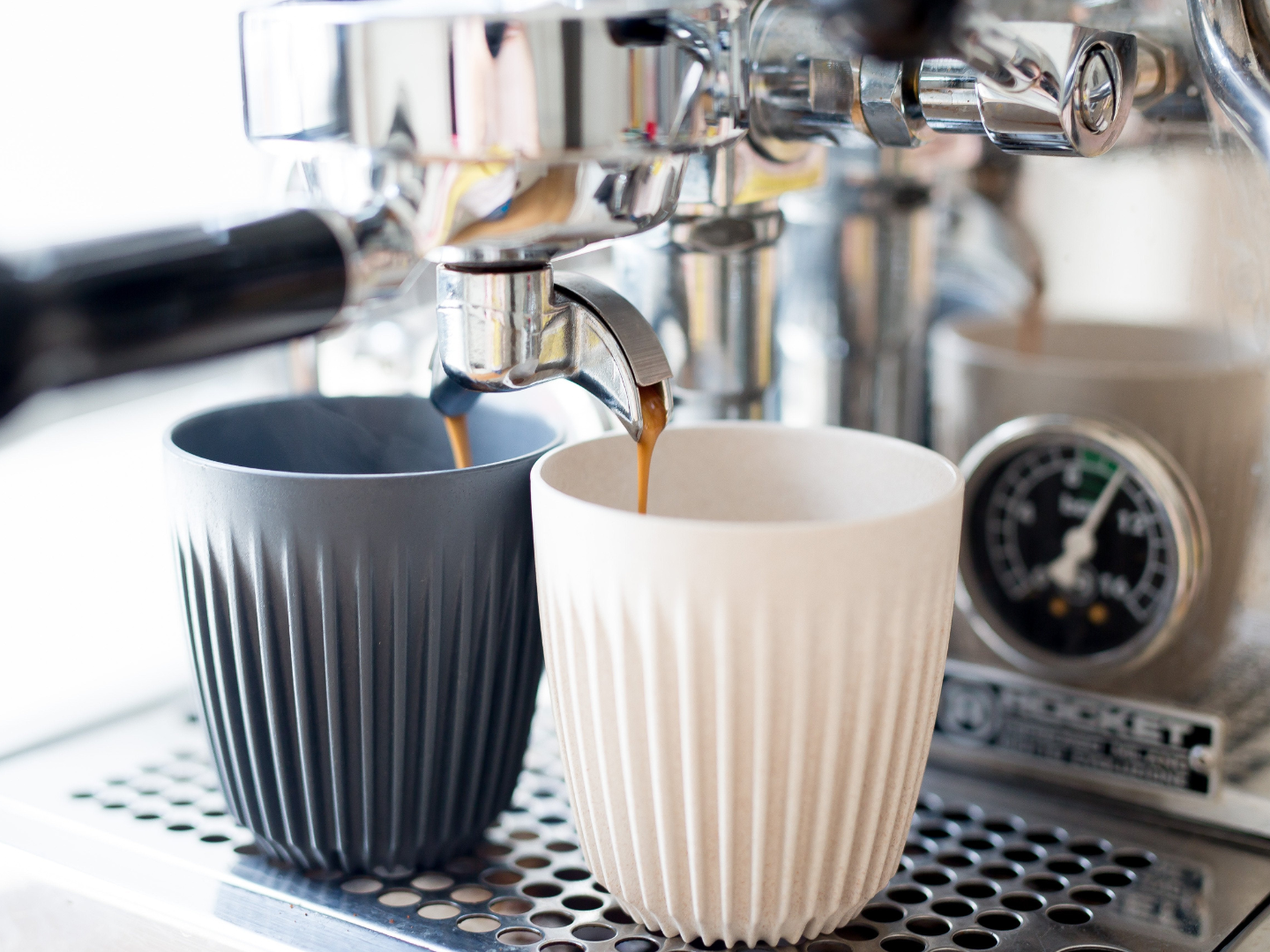 A set of two espresso cups by Huskee placed under a coffee machine that is pouring coffee into them.