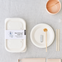 A kintsugi repair kit box laid out on a grey surface, including a white box with tools, gold powder, a paintbrush, gloves, and sticks.