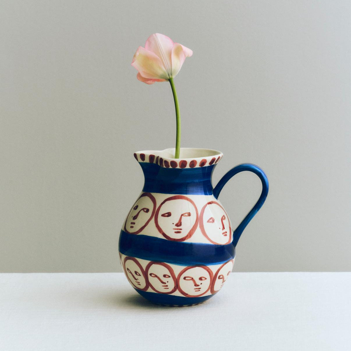Ceramic pitcher with face patterns holding a single pink flower against a plain background