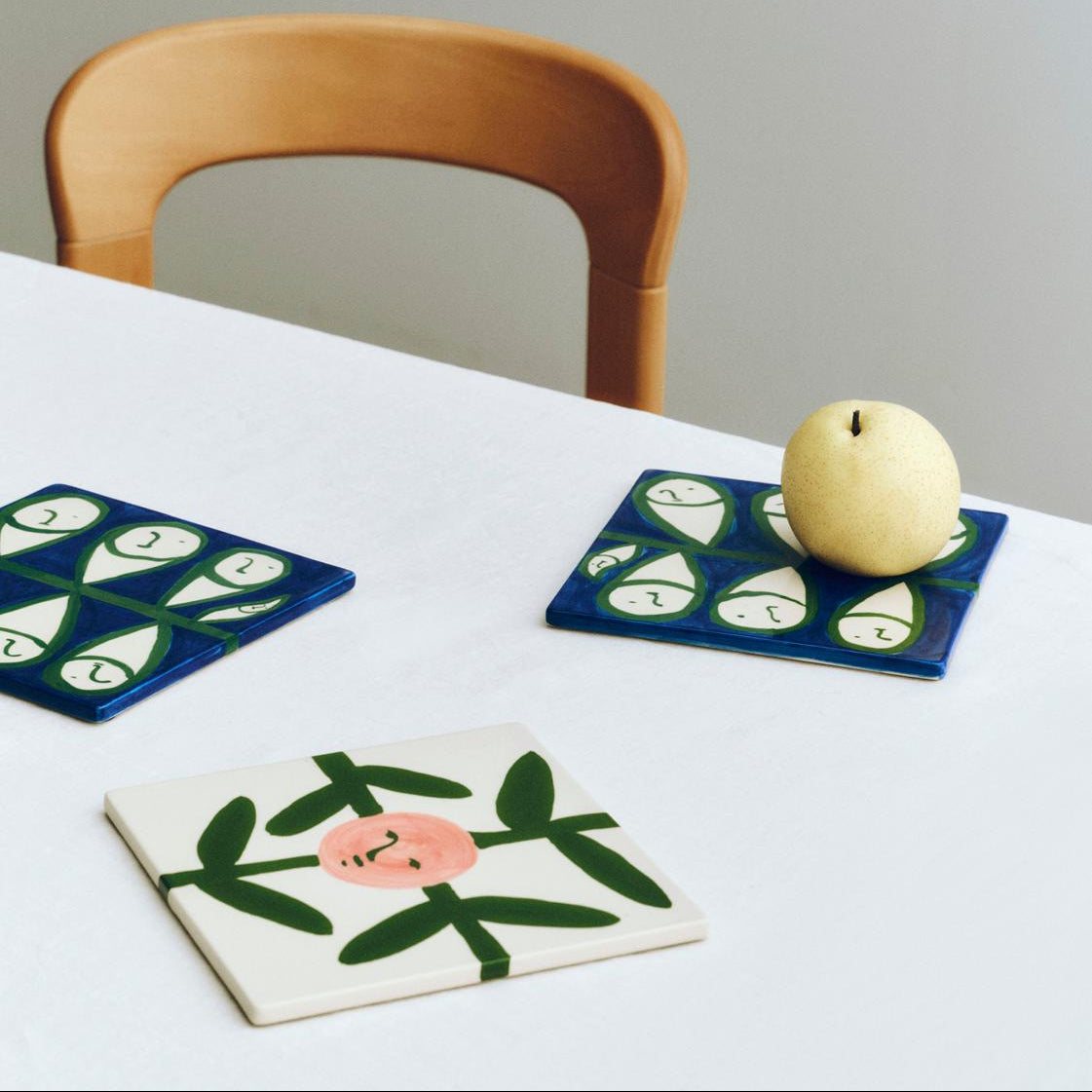 Three ceramic coasters with artistic designs on a white table with a wooden chair.