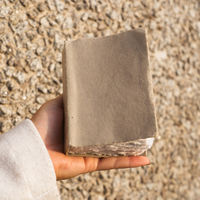 A hand holding a small, beige handmade paper notebook with a solid beige colour cover and a rough deckle edge finish.