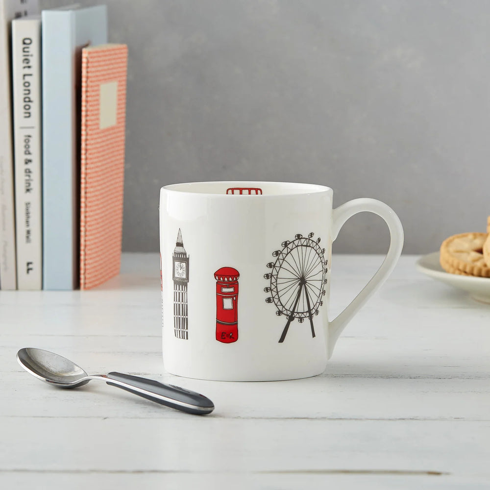 A white mug designed with illustrations of London landmarks and a silver spoon placed next to it