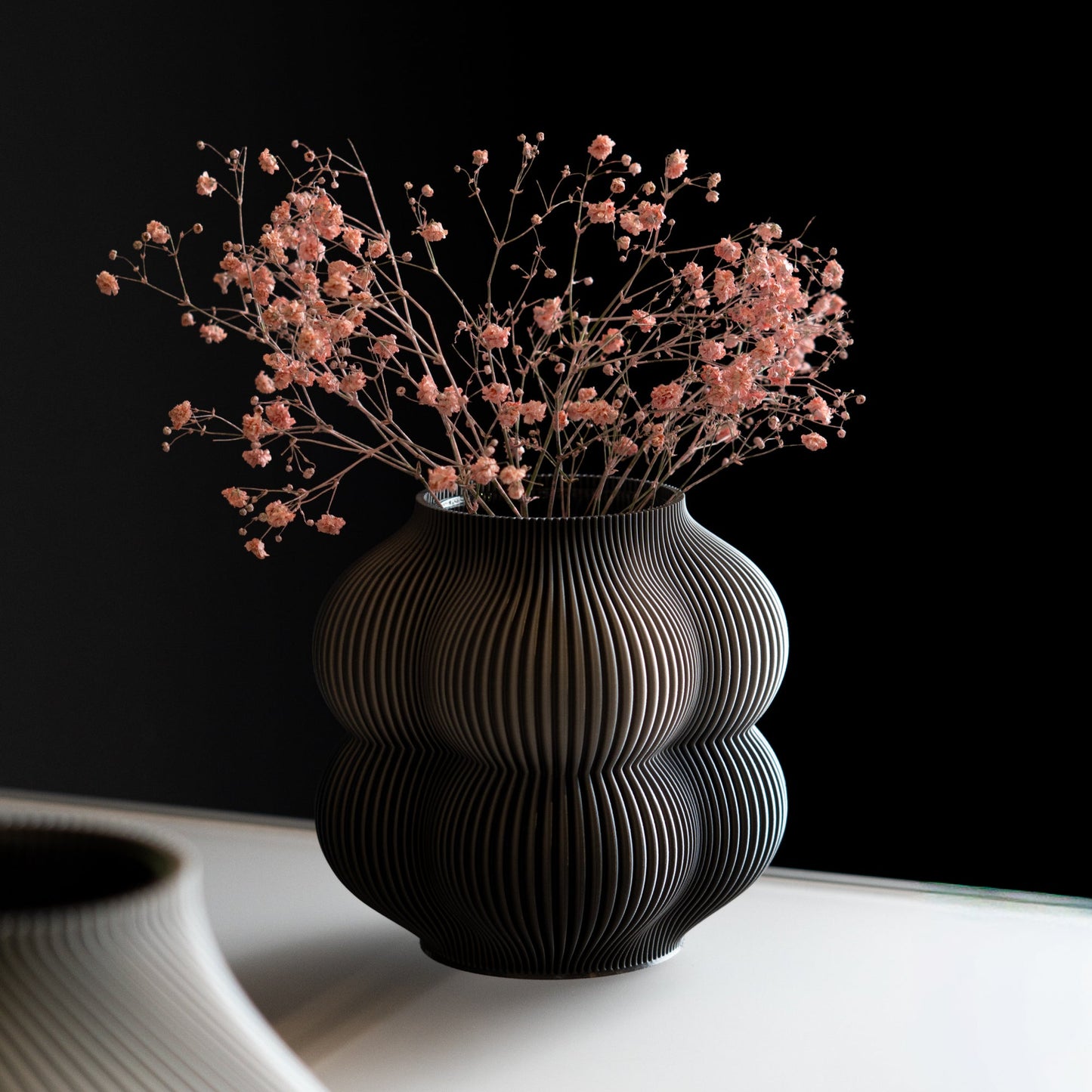 Black ribbed vase with pink dried flowers on a white surface against a black background
