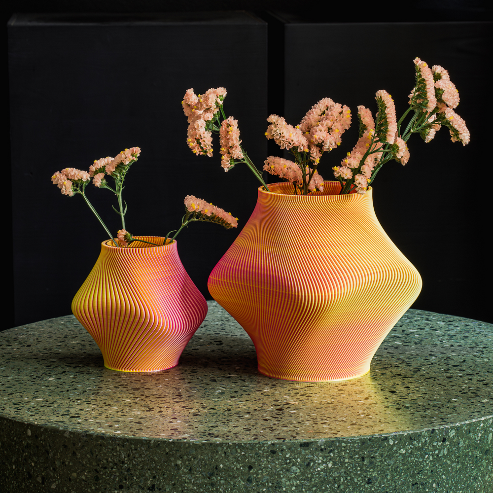 
                  
                    Two orange 3D printed short vases filled with flowers, placed on a stone round table.
                  
                