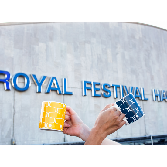 Two hands each holding the net & ball pattern mug in front of the Royal Festival Hall building